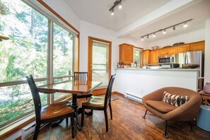 Eine moderne Küche mit Holzausstattung, ein Essbereich mit Tisch und Stühlen sowie große Fenster mit Blick ins Freie.