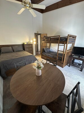 Ein Schlafzimmer mit einem Etagenbett, einem Einzelbett, einem runden Holztisch mit Blumen in einer Vase, einem Deckenventilator und einem Fernseher.