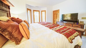 Une chambre à coucher avec un grand lit, un téléviseur posé sur un meuble en bois et une fenêtre équipée de stores.