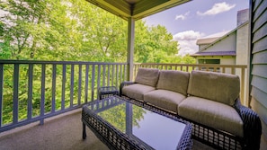 A covered outdoor seating area with a sofa and a glass-top table, surrounded by a wooden railing, overlooking a lush green landscape. - Adirondacks 6 | Fireplace, Restaurant, Pool, Spa (Blowing Rock)