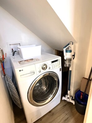 A compact laundry area with a front-loading washing machine, a drying rack, and a vacuum cleaner.