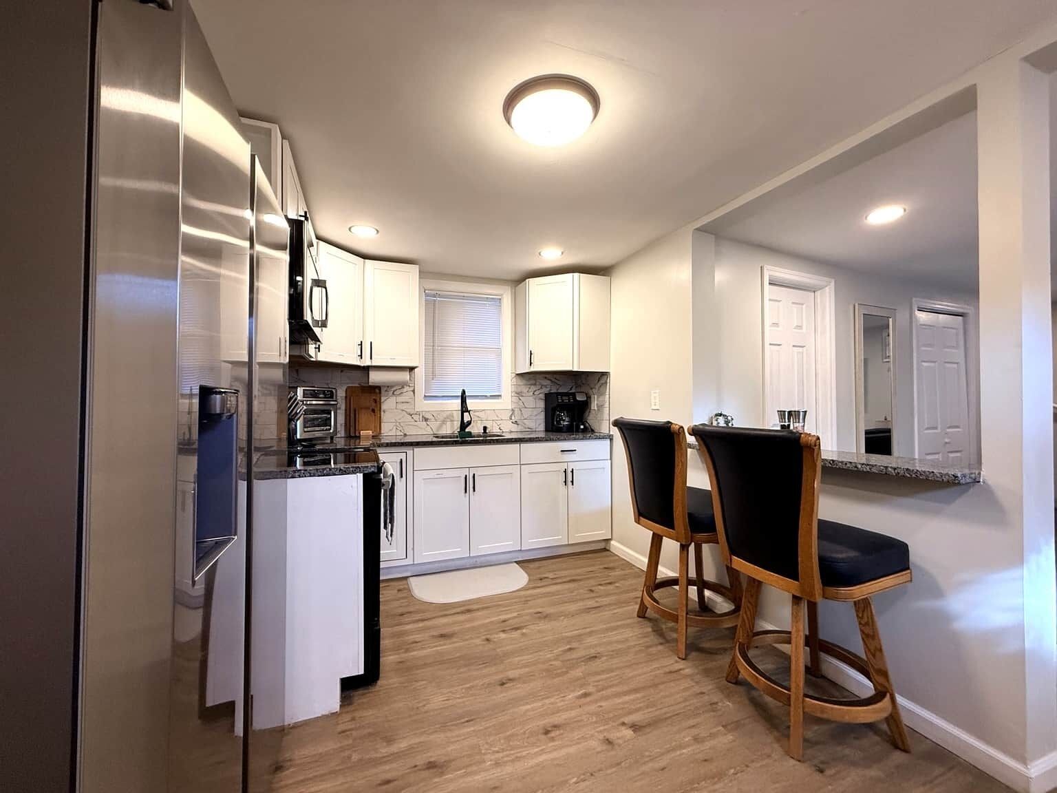A modern kitchen with white cabinets, a built-in oven, and a coffee maker. There are two wooden bar stools and a small island with a sink.