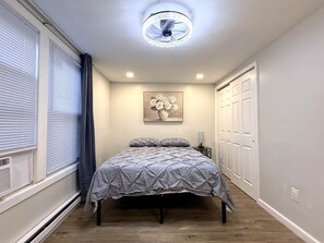 A bedroom with a bed, a ceiling fan, a wall-mounted light, a window with blinds, and a door.