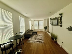 Ein modernes Wohnzimmer mit einer Couch, einem Esstisch und Stühlen. Große Fenster mit Jalousien, ein flachbildfernseher an der Wand und ein Topfpflanze sind vorhanden.