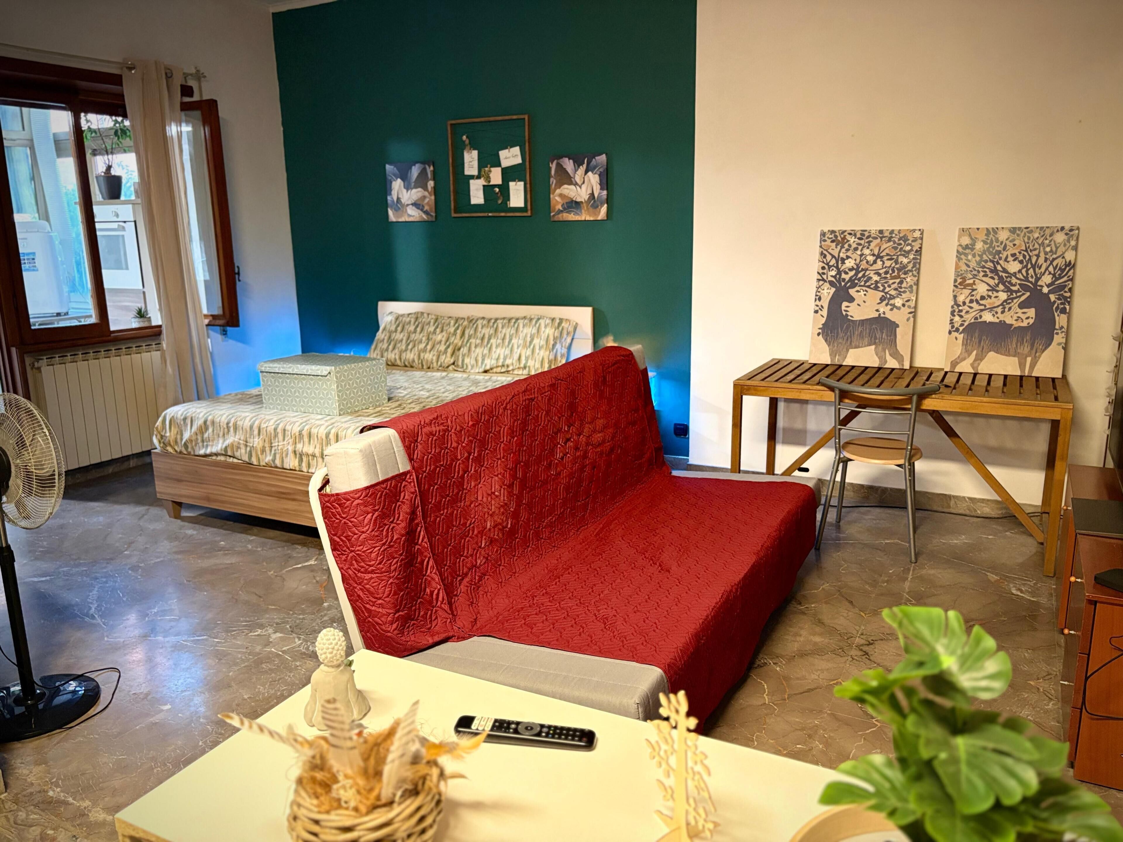 A bedroom with a bed, a red sofa, a small table with a remote control, and artwork on the walls.