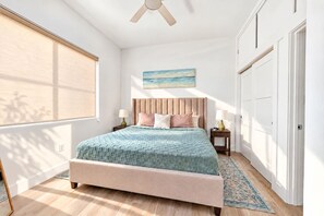 Ein Schlafzimmer mit einem Bett, Nachttischen, einem Deckenventilator, einem Fenster mit Jalousien, einem Gemälde an der Wand und einer Tür.