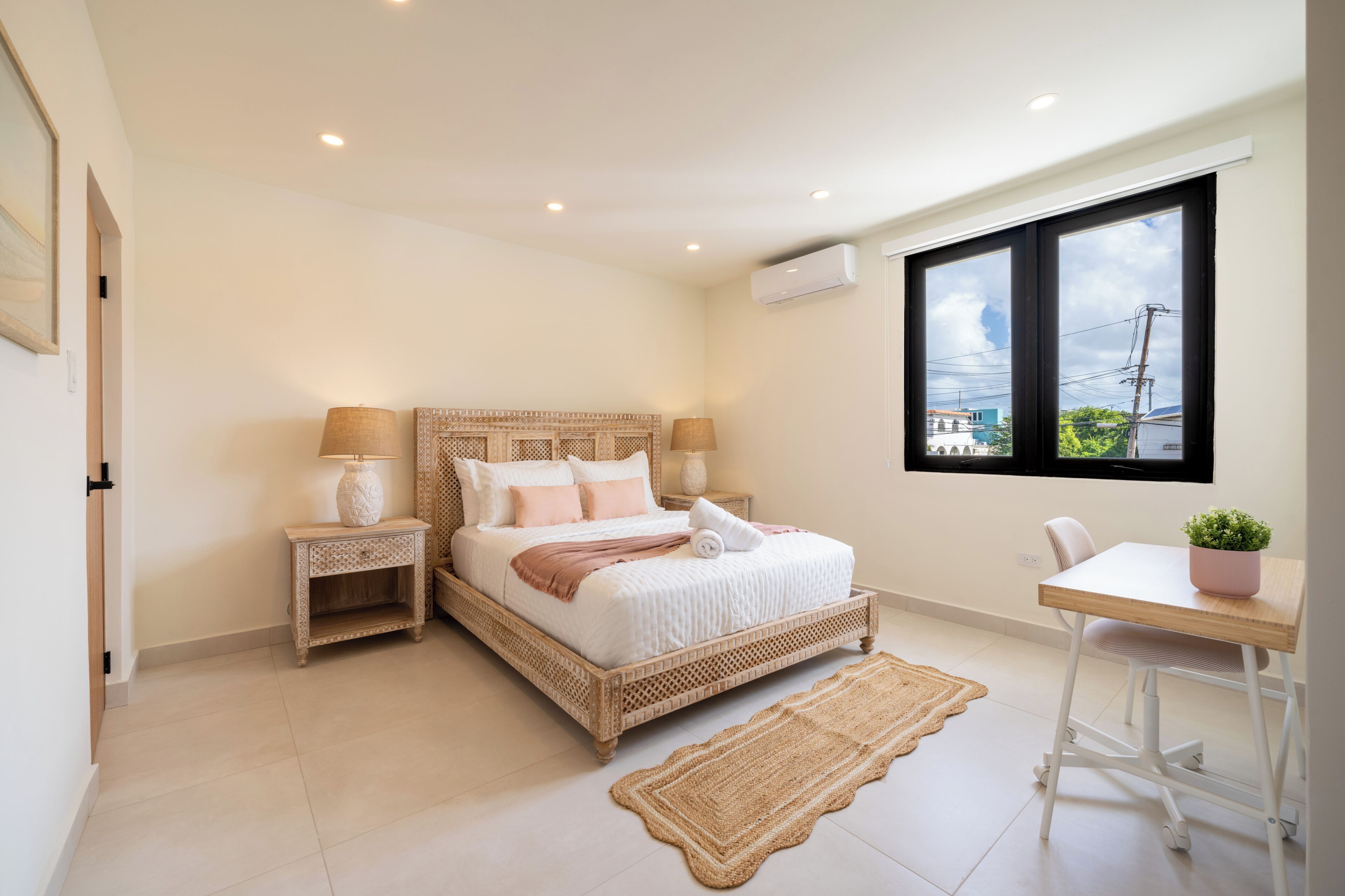 A bedroom with a bed, bedside tables, a chair, a window, and an air conditioning unit.