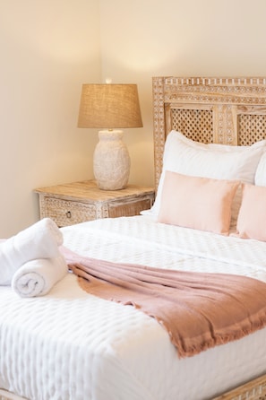 Una cama bien hecha con una manta blanca y rosa, una toalla enrollada y una lámpara sobre una mesita de noche de madera.