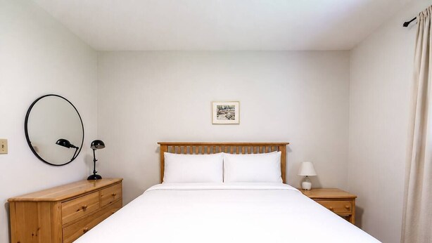 Un dormitorio con cabecera de madera, una cama individual, una mesita de noche con lámpara, un aparador y un cuadro en la pared.