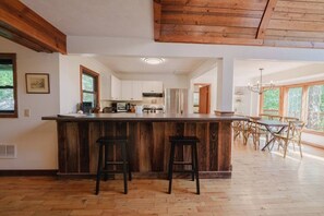 Eine geräumige Küche mit einer Holztischinsel und Barhocker, ein Essbereich mit Tisch und Stühlen sowie große Fenster, die für natürliches Licht sorgen.