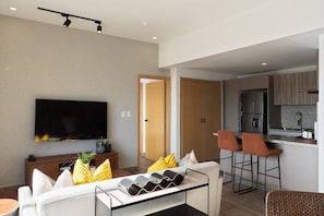 A modern living room with a flat-screen TV, a sofa, and a coffee table. There is a kitchen area with a refrigerator and bar stools. - Amazing Apt W/ Pool In Regatta Living By YellowKey (Santo Domingo)