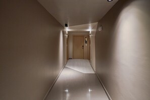 Un estrecho pasillo bien iluminado con una puerta de madera al final, suelo de baldosas e interruptor de luz en la pared.