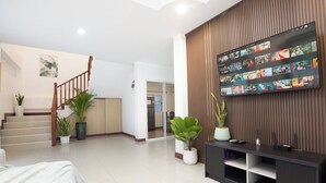 A modern living room with a flat-screen TV displaying various shows, a staircase, and potted plants. - 4BR Family Home • Garden & Sala • Sleeps 9 • Near Airport (Nong Phueng)