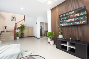 A modern living room with a flat-screen TV displaying various shows, a staircase, and potted plants. - 4BR Family Home • Garden & Sala • Sleeps 9 • Near Airport (Nong Phueng)