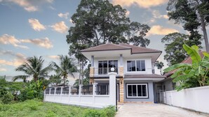 A two-story house with a balcony, surrounded by greenery and trees. - 4BR Family Home • Garden & Sala • Sleeps 9 • Near Airport (Nong Phueng)