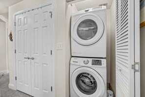 A laundry area with a front-loading washer and dryer, a white door with a handle, and a small shelf with items.