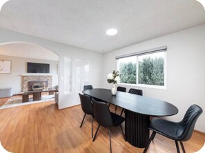 A modern living room with a fireplace, a dining table with chairs, and a large window with a view of trees.