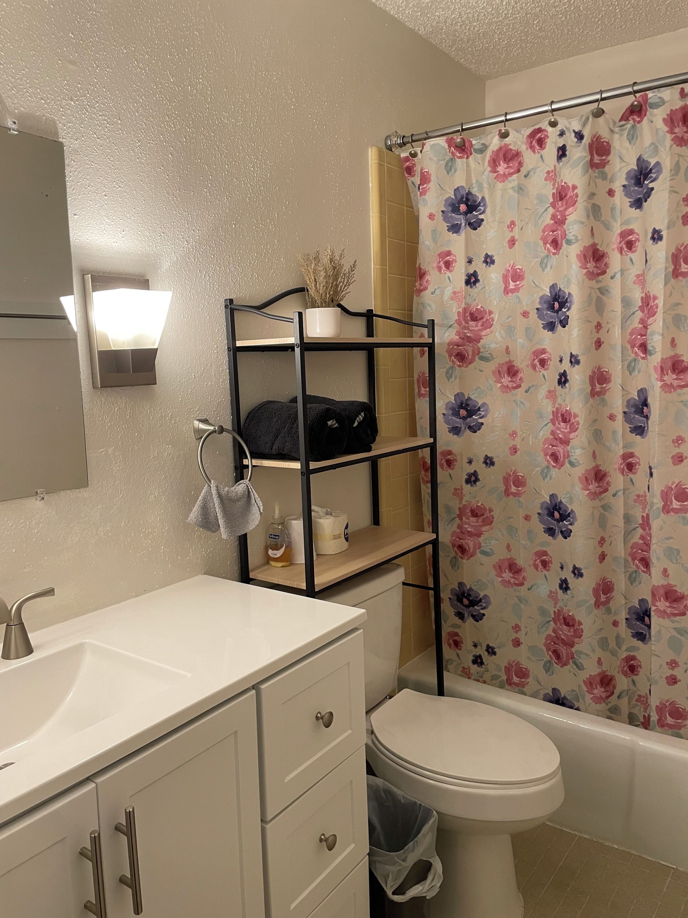 Un baño con un lavabo blanco, un inodoro, una cortina de ducha con estampado floral y un estante de metal negro con toallas y artículos de aseo.