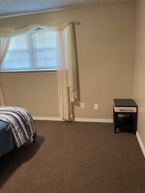 A bedroom with a bed, a nightstand, a window with blinds, and a curtain.