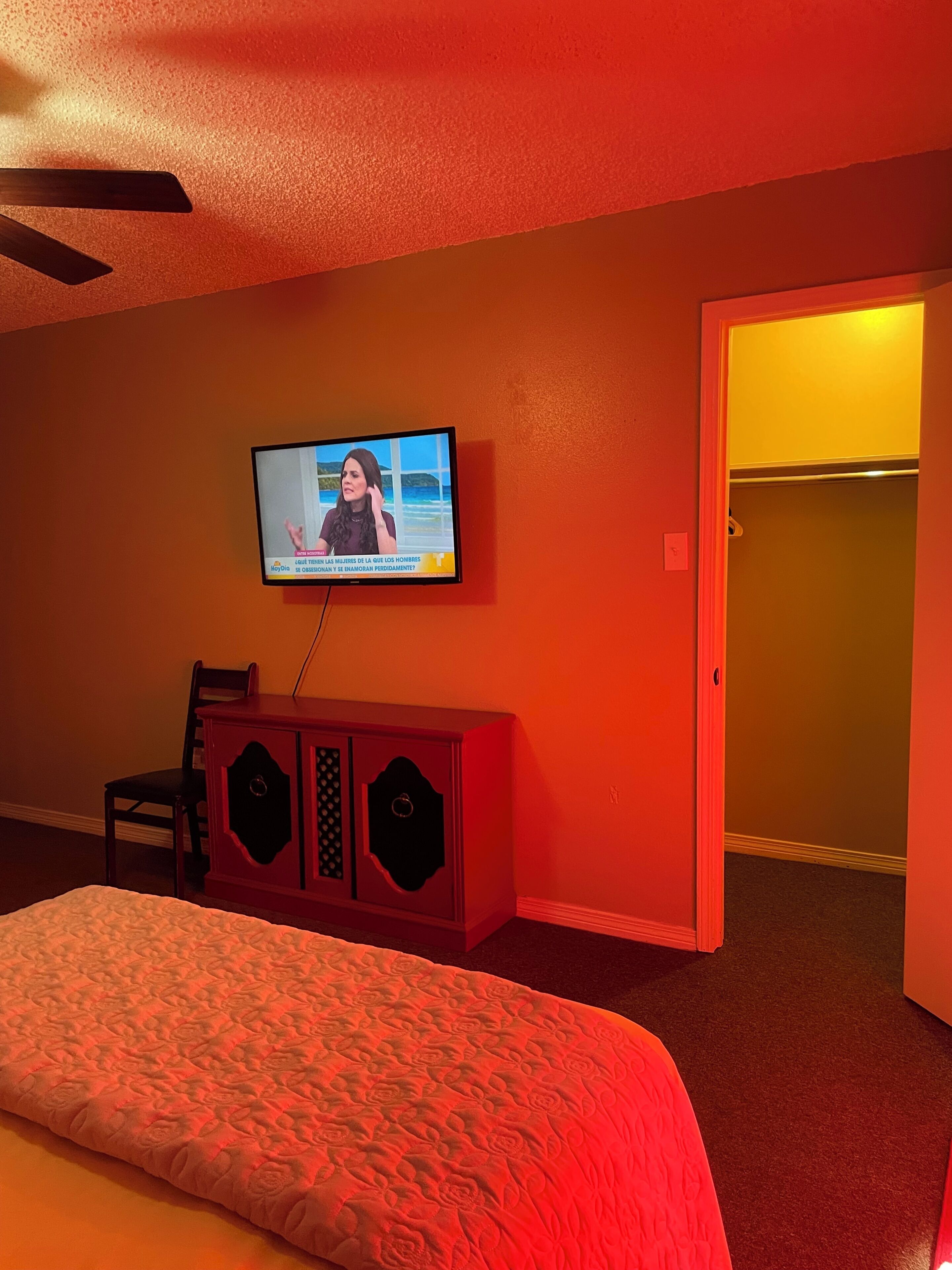Habitación de hotel con televisión montada en la pared, cama con colcha estampada, un armario rojo y una puerta que da a otra habitación.