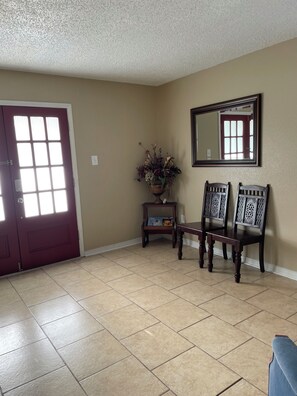A room with a red door, a mirror, two chairs, a small table with a vase, and beige walls.