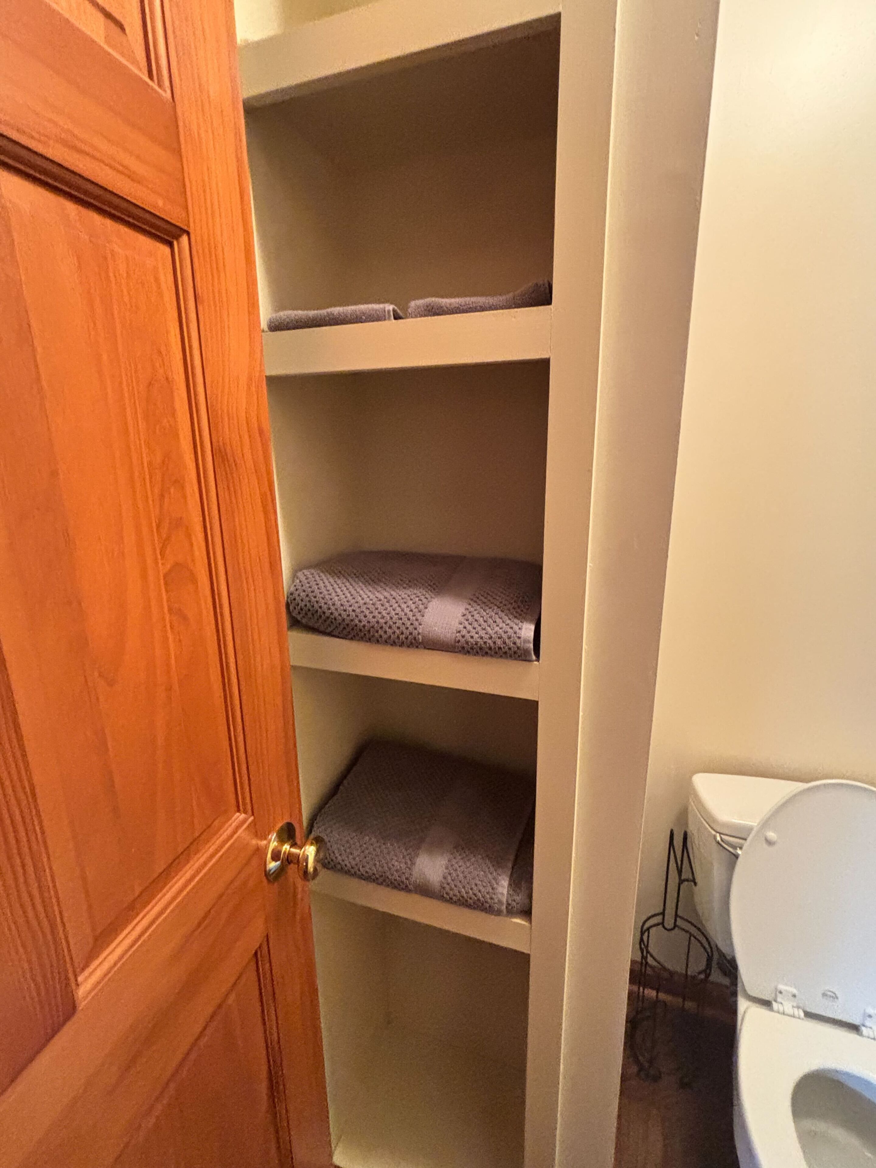 A bathroom with a toilet, a wooden door, and a shelf unit with folded towels.