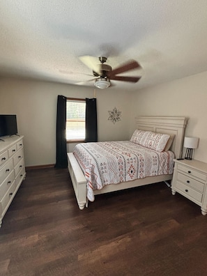 A bedroom with a bed, dresser, nightstand, ceiling fan, and a decorative wall piece.