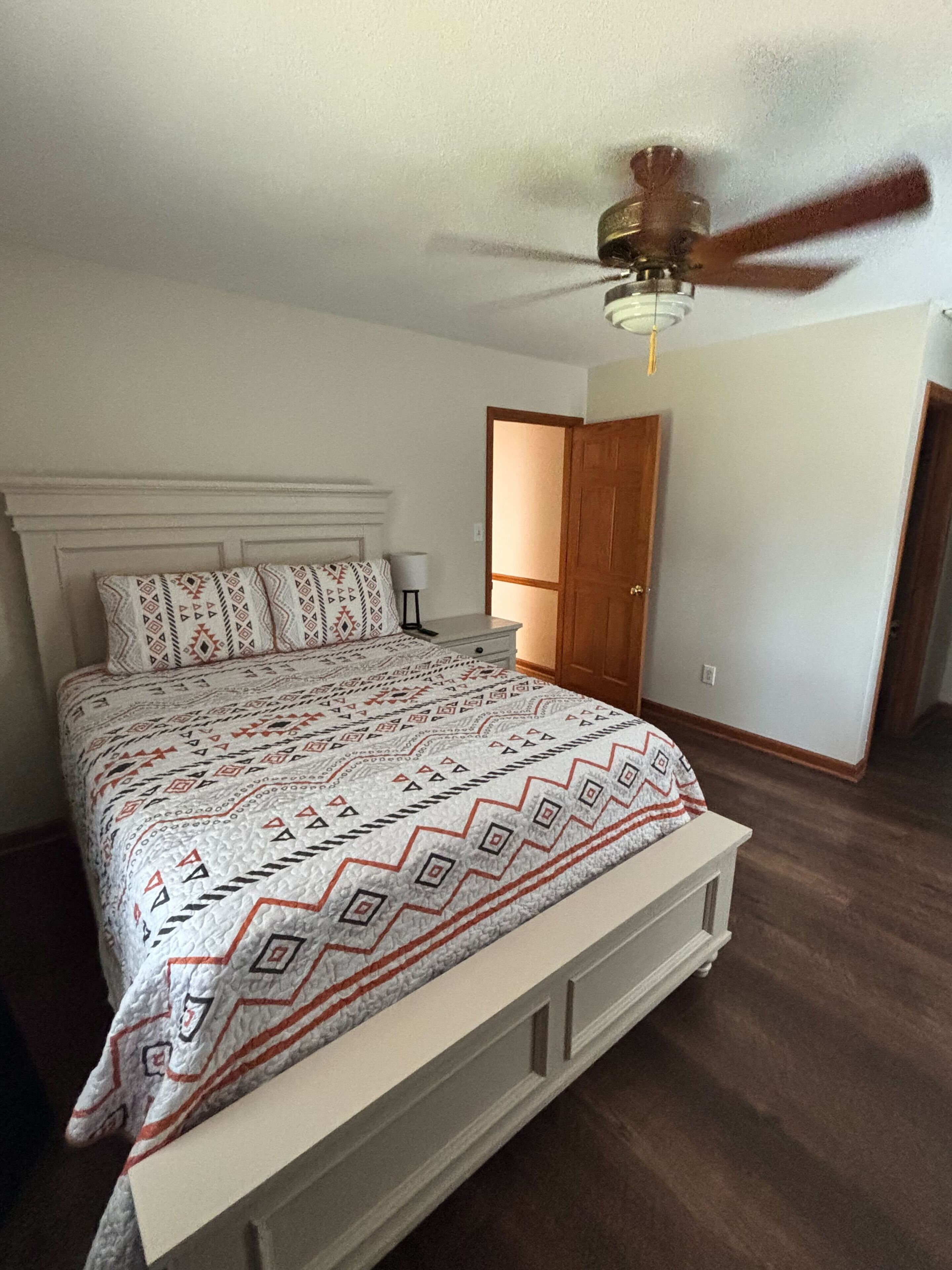 A bedroom with a bed, a ceiling fan, a door, and a nightstand.