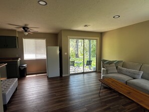 A living room with a sofa, a coffee table, a kitchen area with a refrigerator, and a sliding glass door leading to an outdoor patio.