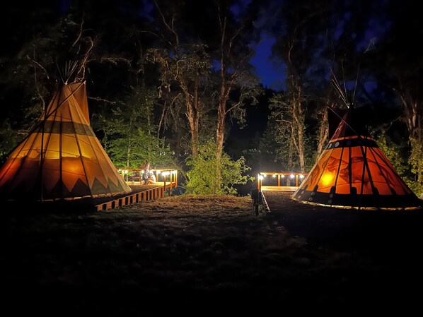 Two illuminated teepees with a wooden deck in between, set in a forest at night.