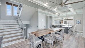 A modern kitchen with a central island, a dining area with a wooden table and wicker chairs, and a staircase with a railing. - Singing Waves (Port St. Joe)