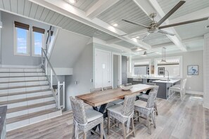A modern kitchen with a central island, a dining area with a wooden table and wicker chairs, and a staircase with a railing. - Singing Waves (Port St. Joe)