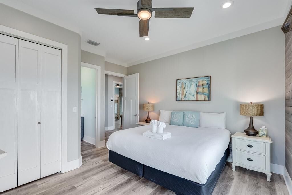 A bedroom with a bed, a ceiling fan, a nightstand with a lamp, and a painting on the wall.