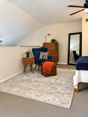 A bedroom with a sloped ceiling, a ceiling fan, a wooden dresser, a blue armchair, a small table with a lamp, a mirror, and a patterned rug. - Modern Villa+ Scottsdale Resort + Pool + Golf Course+ Walking paths +Gym +Dining (Scottsdale)