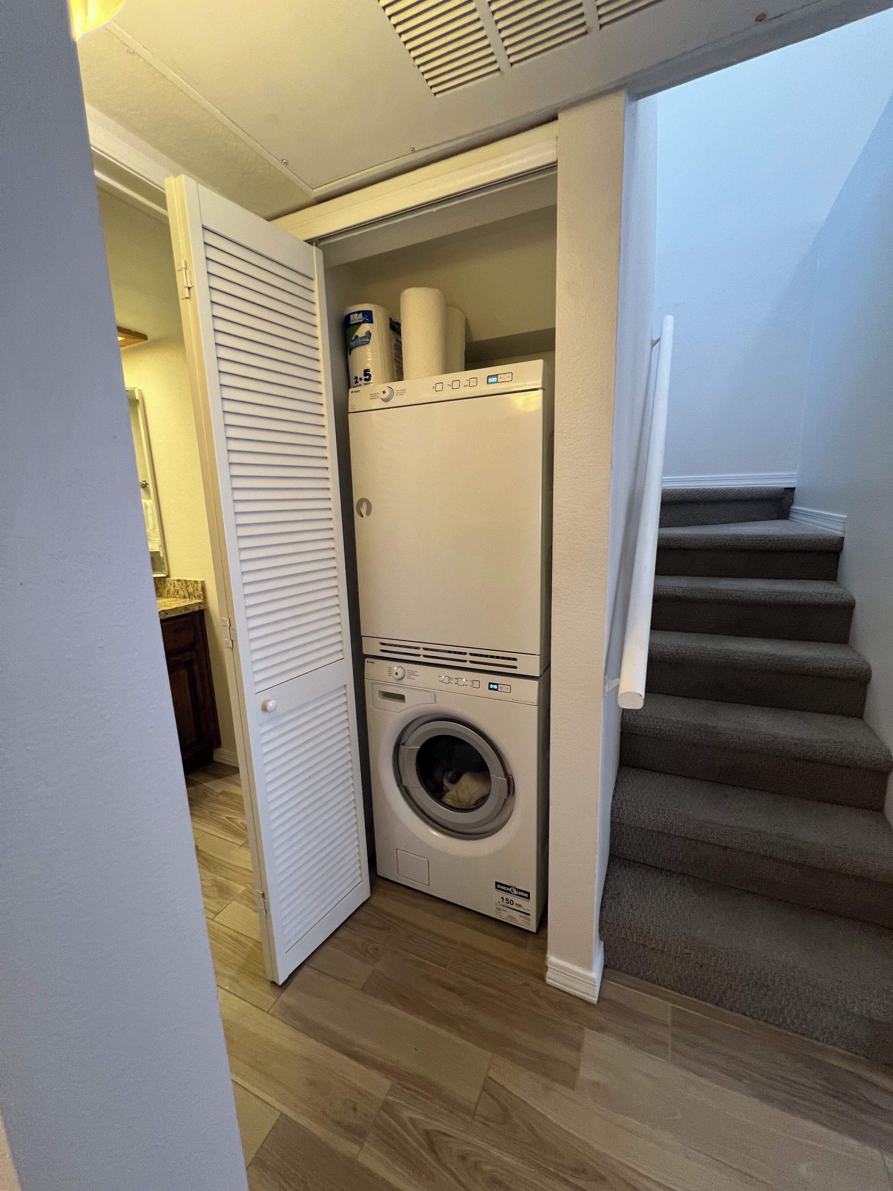 A compact laundry room with a front-loading washer and dryer, a wooden floor, and a staircase leading to an upper level.
