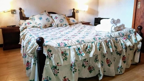 Quarto com colcha florida, cabeceira de madeira, luminárias de cabeceira e piso de madeira.
