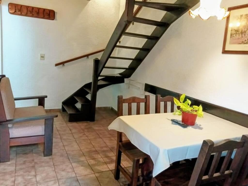 Un área de comedor con una mesa, sillas, una planta en maceta, una escalera y un cartel en la pared.