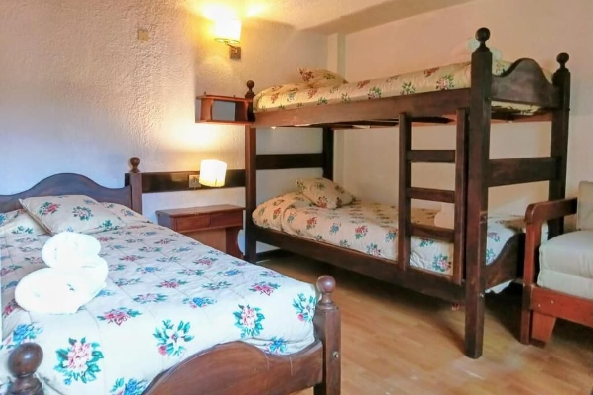 Una habitación con una cama litera, una cama individual y una mesita de noche con lámpara.