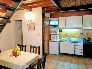 Cocina compacta con microondas, fregadero y estufa. Hay una mesa de comedor con sillas y una planta en maceta. Un televisor está montado en la pared.
