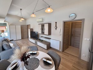 A modern living room with a sofa, dining table, and a TV. There are pendant lights, a clock, and a door. - Penthouse with a Panorama balcony, and free Parking! (Kecskemét)