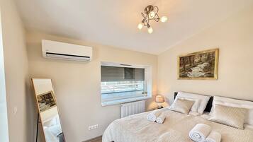 A bedroom with a bed, a window, a painting, an air conditioner, and a mirror.