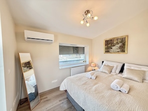 A bedroom with a bed, a window, a painting, an air conditioner, and a mirror.