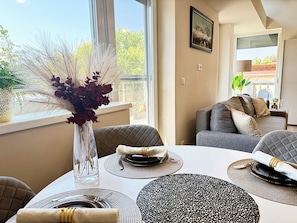 A dining area with a table set for four, a vase with dried plants, and a view of the outdoors through a window.