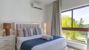 Et hotelværelse med seng, natbord, lampe, aircondition og et vindue med udsigt til palmer.