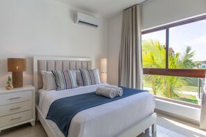Ein Hotelzimmer mit Bett, Nachttisch, Lampe, Klimaanlage und einem Fenster mit Blick auf Palmen.