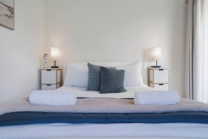 A neatly made bed with two pillows, a bedside table with a lamp, and a window with curtains. - Willow Landing Beach House with Private Jetty (Frankston)