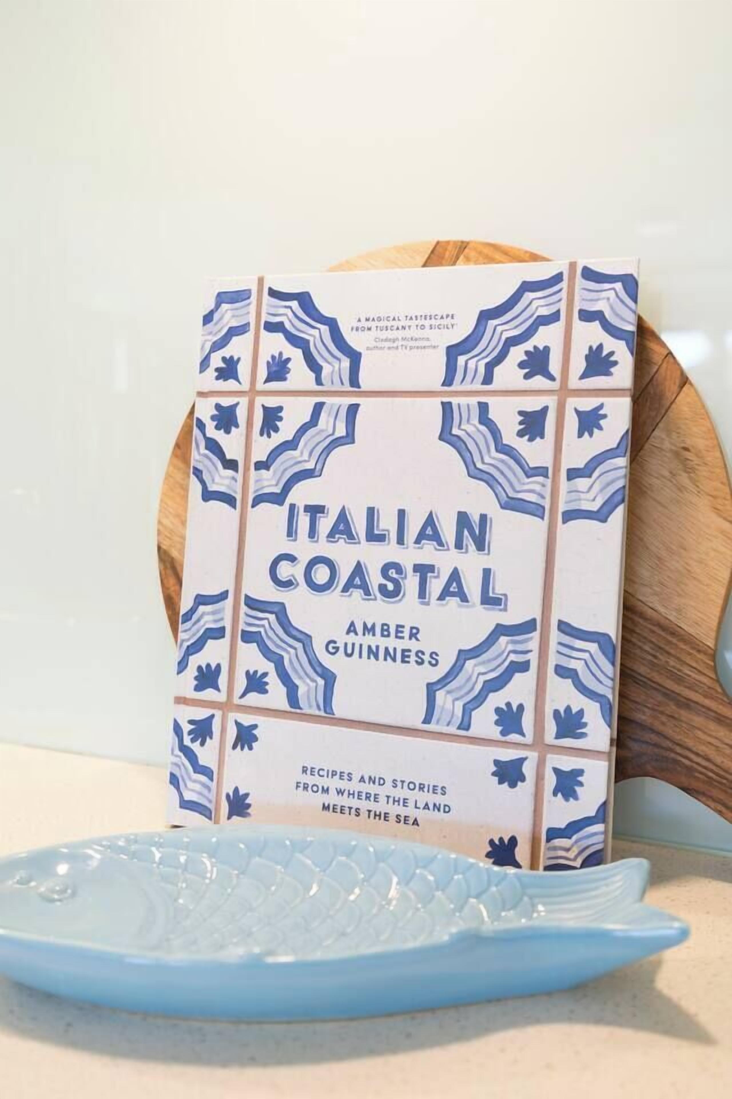 Ein Buch mit dem Titel 'Italian Coastal' von Amber Guinness, platziert auf einer Holzplatte, neben der ein blauer Teller steht.