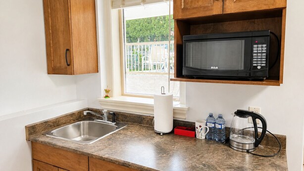 A kitchen with a stainless steel sink, wooden cabinets, a microwave, a kettle, and a window with blinds.