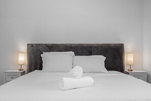 Ein Bett mit weißen Laken, zwei Kissen, ein aufgerolltes Handtuch und zwei Nachttischlampen auf Nachttischen.