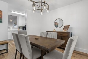 A dining area with a wooden table and grey upholstered chairs, a chandelier, and a kitchen in the background. - Modern Getaway & Casita Near the Domain + Fire Pit Backyard, Sleeps 8 (Austin)